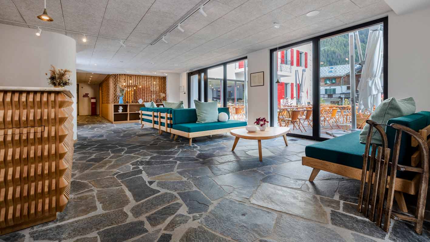 Espace lounge lumineux de l'hôtel Couronne à Chamonix, avec des canapés en bois et coussins verts sur mesure, une table basse en bois clair et un sol en pierre naturelle, offrant une ambiance chaleureuse et contemporaine