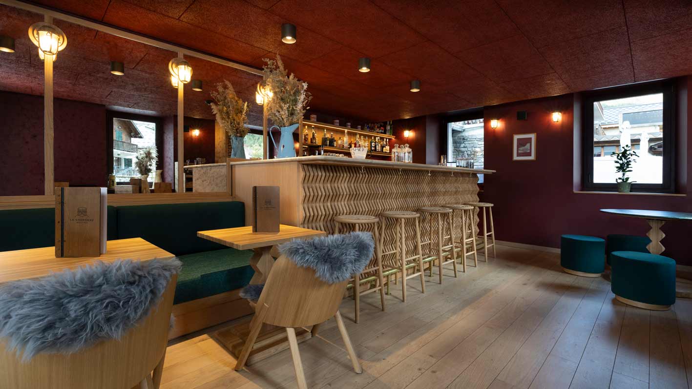 Espace bar de l'hôtel la Couronne à Chamonix avec une peinture rouge aux murs et plafonds, chaises en bois, tables en bois et banquette en bois et tissu bleu, et un bar en bois sculpté, le tout dans un esprit chaleureux type montagne
